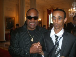A photo of Stevie Wonder and Mussie Gebre. Both men in dark suits: Stevie is wearing a pin striped dark suit, black t-shirt, and has a gold pendant; Mussie is wearing a dark suit, white shirt, and a striped black/white tie. The men are holding hands.